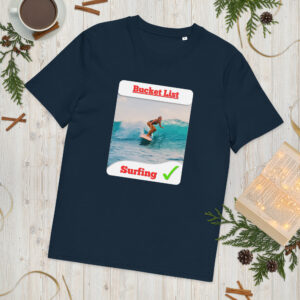 Navy T-shirt laid flat with a poster reading Bucket List showing a surfer, surrounded by winter greenery and wooden surface elements.